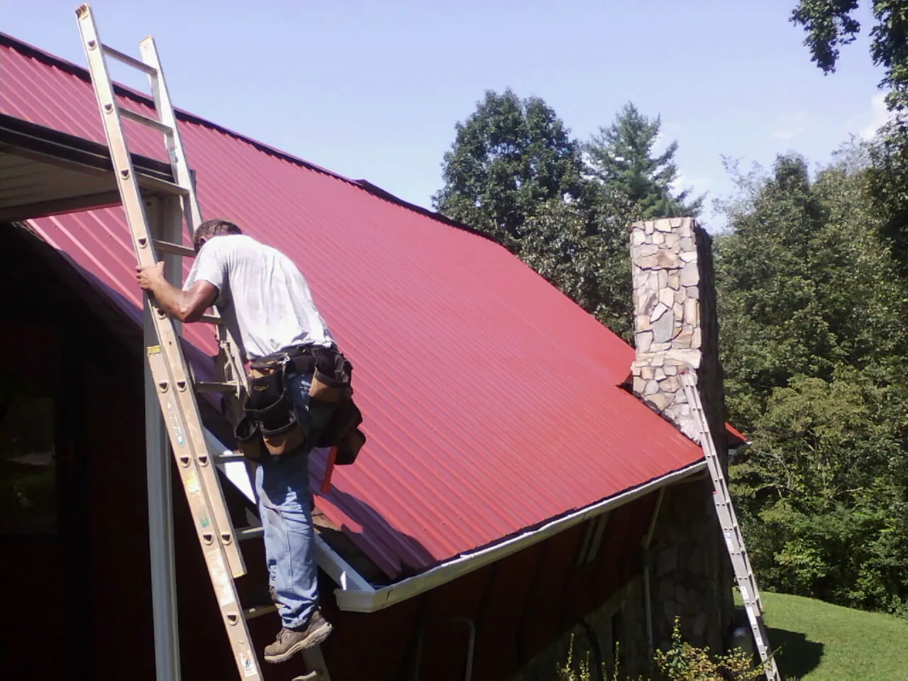 Roofer on a ladder beside a red metal roof and stone chimney — Soffit & Fascia Repair by Roofing Brothers in Alexander City